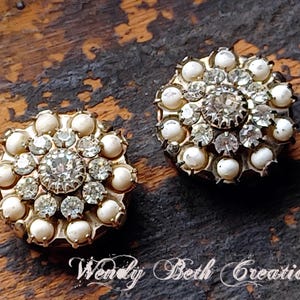 Pode incluir: Dois botões redondos de estilo vintage com base em metal dourado. Cada botão apresenta um grande strass transparente central rodeado por strass transparentes menores e pérolas de imitação creme. Os botões estão sobre um fundo escuro e texturizado.