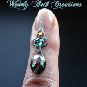 May include: A silver charm with a teal, iridescent, and yellow gemstone design. The charm is hanging from a finger.
