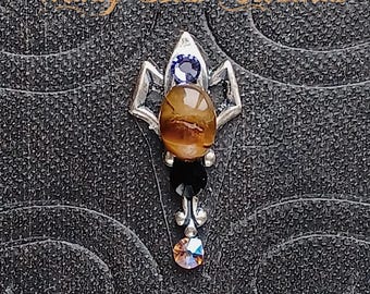 May include: A silver pendant with a tiger's eye stone, a purple crystal, and a black crystal. The pendant is in the shape of a flower.