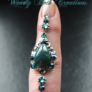 May include: A finger adorned with a silver ring featuring a teardrop-shaped green gemstone surrounded by smaller rhinestones. The ring is accented with smaller green and black rhinestones on the finger.