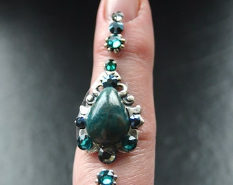 May include: A finger adorned with a silver ring featuring a teardrop-shaped green gemstone surrounded by smaller rhinestones. The ring is accented with smaller green and black rhinestones on the finger.