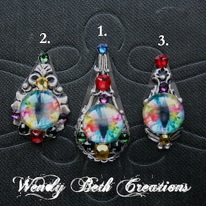May include: Three ornate, teardrop-shaped pendants with iridescent, eye-shaped cabochons. Each pendant features a silver-toned metal setting adorned with colourful gemstones. The text "Wendy Beth Creations" is at the bottom.