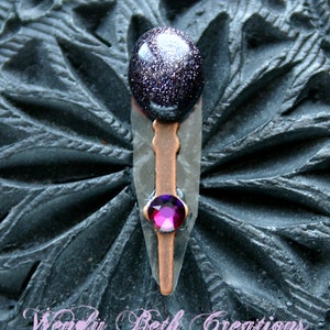 May include: A handcrafted pendant featuring a dark blue, speckled stone cabochon at the top, a copper-colored metal accent, and a vibrant pink gemstone at the bottom. The pendant is set against a dark, textured background. The text "Wendy Beth Creations" is at the bottom.
