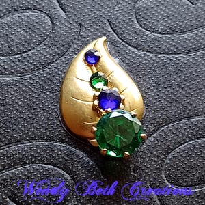 May include: A gold-toned leaf-shaped brooch with a series of colorful gemstones. The brooch features a large, round, emerald-green stone at the base, with smaller blue and green stones ascending the leaf. The brooch has the text "Wendy Beth Creations".