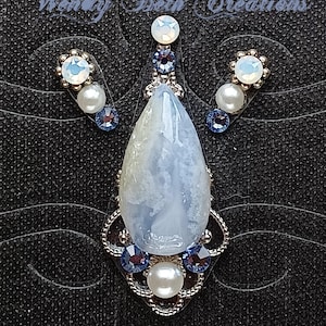 May include: A jewellery set comprising a pendant and earrings. The pendant features a large, teardrop-shaped, pale blue stone with white marbling, set in silver filigree with blue and white accents. The earrings echo the design with smaller stones.
