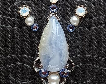 May include: A jewelry set featuring a pendant and matching earrings. The pendant has a large, teardrop-shaped, pale blue stone with white marbling, surrounded by silver filigree and blue and white accents. The earrings mirror the design with smaller stones.