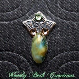 Może przedstawiać: Srebrny i zielony metalowy urok z zielonym kamieniem szlachetnym i przezroczystym owalnym kaboszonem. Urok ma kształt gwiazdy z ozdobnym wzorem.