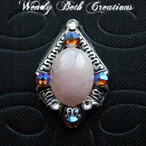 May include: A silver-toned pendant with a large, oval, light pink stone at its center. The pendant is adorned with smaller, iridescent crystals in shades of blue, orange, and clear. The text "Wendy Beth Creations" is at the top.