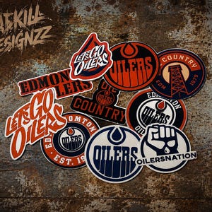 May include: A collection of Edmonton Oilers themed vinyl stickers featuring various team logos, text including Let's Go Oilers, Oil Country, and Oilersnation, rendered in orange, navy blue, and white colours, arranged in a cluster on a textured surface.