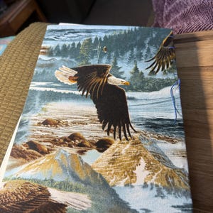K&ouml;nnte beinhalten: Ein stoffbezogenes Notizbuch mit einer detaillierten Illustration von Wei&szlig;kopfseeadlern, die &uuml;ber einer Berglandschaft fliegen. Das Design umfasst Blau-, Braun- und Wei&szlig;t&ouml;ne und hebt die Fl&uuml;gel der Adler und die Naturlandschaft hervor.