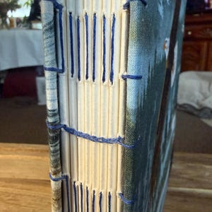 K&ouml;nnte beinhalten: Ein handgefertigtes Notizbuch mit einem blau-wei&szlig; gemusterten Einband und cremefarbenen Seiten. Der R&uuml;cken ist mit blauem Faden handgen&auml;ht. Das Cover zeigt eine Naturszene mit B&auml;umen und blauem Himmel.