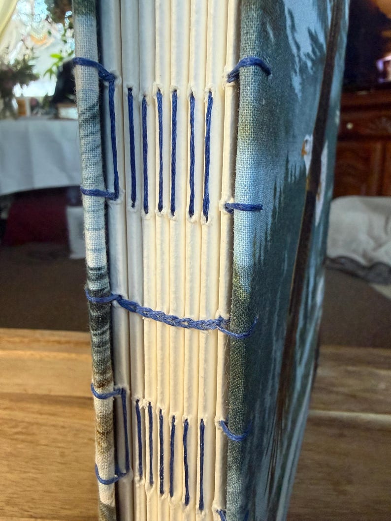K&ouml;nnte beinhalten: Ein handgefertigtes Notizbuch mit einem blau-wei&szlig; gemusterten Einband und cremefarbenen Seiten. Der R&uuml;cken ist mit blauem Faden handgen&auml;ht. Das Cover zeigt eine Naturszene mit B&auml;umen und blauem Himmel.