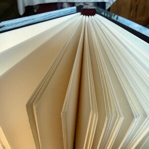 K&ouml;nnte beinhalten: Offenes Buch mit cremefarbenen, aufgef&auml;cherten Seiten. Die Seiten sind dick und strukturiert, mit einem leichten Off-White-Farbton. Der Einband ist sichtbar, mit einem dunkelblauen und schwarzen Einband.