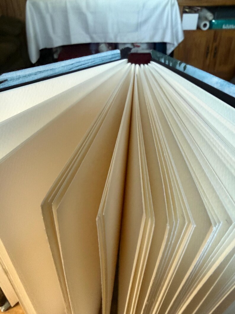 K&ouml;nnte beinhalten: Offenes Buch mit cremefarbenen, aufgef&auml;cherten Seiten. Die Seiten sind dick und strukturiert, mit einem leichten Off-White-Farbton. Der Einband ist sichtbar, mit einem dunkelblauen und schwarzen Einband.