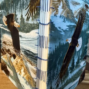 K&ouml;nnte beinhalten: Ein handgefertigtes Notizbuch mit einem Stoffeinband, der einen majest&auml;tischen Wei&szlig;kopfseeadler zeigt, der &uuml;ber einer Berglandschaft schwebt. Die Seiten sind mit blauem Faden handgen&auml;ht.