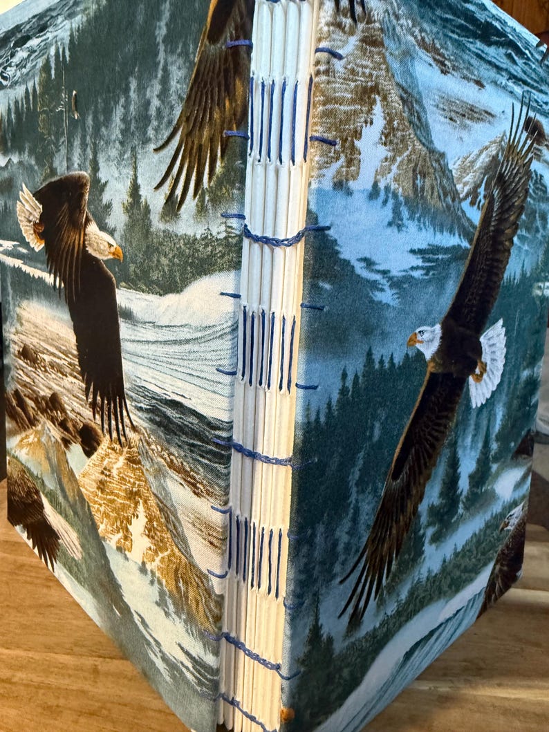 K&ouml;nnte beinhalten: Ein handgefertigtes Notizbuch mit einem Stoffeinband, der einen majest&auml;tischen Wei&szlig;kopfseeadler zeigt, der &uuml;ber einer Berglandschaft schwebt. Die Seiten sind mit blauem Faden handgen&auml;ht.