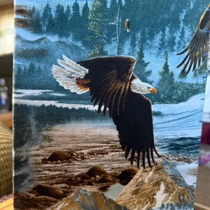 K&ouml;nnte beinhalten: Stoffpaneel mit einem majest&auml;tischen Wei&szlig;kopfseeadler im Flug &uuml;ber einer Bergkette und einem Fluss. Der Adler hat braune und wei&szlig;e Federn mit einem gelben Schnabel. Der Hintergrund zeigt einen Wald und einen blauen Himmel.