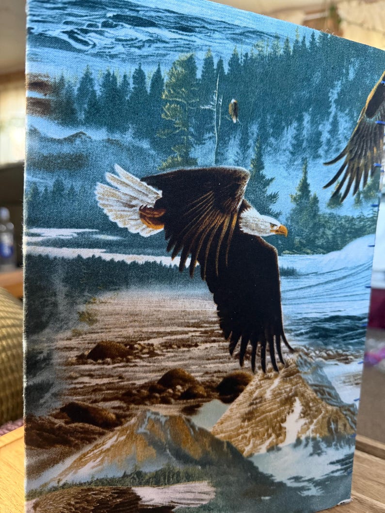K&ouml;nnte beinhalten: Stoffpaneel mit einem majest&auml;tischen Wei&szlig;kopfseeadler im Flug &uuml;ber einer Bergkette und einem Fluss. Der Adler hat braune und wei&szlig;e Federn mit einem gelben Schnabel. Der Hintergrund zeigt einen Wald und einen blauen Himmel.