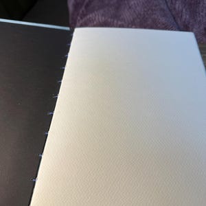 K&ouml;nnte beinhalten: Ein offenes Skizzenbuch mit einem dunkelbraunen Einband und cremefarbenen Seiten. Die Seiten sind mit blauem Faden zusammengen&auml;ht. Das Papier hat eine strukturierte Oberfl&auml;che. Das Skizzenbuch ist ge&ouml;ffnet und zeigt die leeren Seiten.