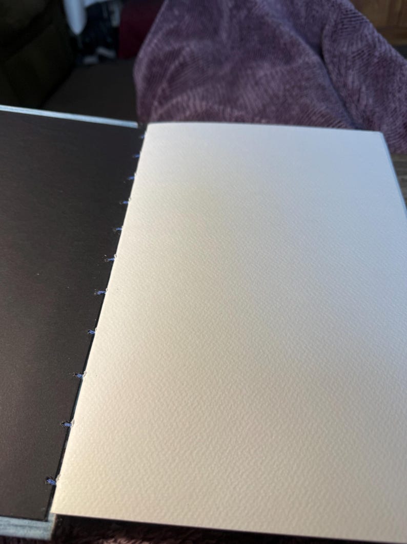K&ouml;nnte beinhalten: Ein offenes Skizzenbuch mit einem dunkelbraunen Einband und cremefarbenen Seiten. Die Seiten sind mit blauem Faden zusammengen&auml;ht. Das Papier hat eine strukturierte Oberfl&auml;che. Das Skizzenbuch ist ge&ouml;ffnet und zeigt die leeren Seiten.