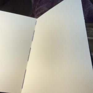 K&ouml;nnte beinhalten: Ein offenes Skizzenbuch mit leeren, cremefarbenen Seiten. Die Seiten werden durch blaue N&auml;hte entlang des R&uuml;ckens zusammengehalten. Das Buch ist ge&ouml;ffnet und zeigt das strukturierte Papier, bereit zum Zeichnen oder Schreiben.