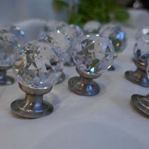 Puede incluir: Un juego de ocho pomos de armario de cristal transparente con superficies facetadas y bases plateadas. Los pomos están dispuestos en semicírculo sobre una superficie blanca.