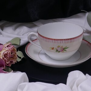 Puede incluir: Una taza de té y un platillo blancos con un diseño floral. La taza de té tiene un borde rojo y una rosa rosa con hojas verdes. El platillo tiene un borde rojo y un borde dorado.