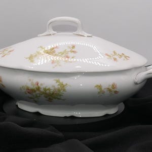 May include: White porcelain serving dish with lid and handles. The dish features a floral pattern in pink and green. The lid has a handle on top. The dish is oval-shaped and sits on a black surface.