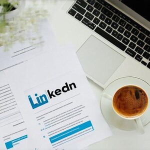 May include: A white laptop with a black keyboard and a cup of coffee on a saucer. A document with the LinkedIn logo is on the table, along with other papers and a small vase of flowers.