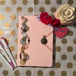 May include: A pink journal adorned with decorative gears and a bronze pocket watch charm. The journal is next to three highlighters and a heart-shaped decoration with the words "BE MINE". Red and cream-coloured flowers are also present.