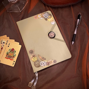 May include: A green journal with a decorative metal clasp and a variety of gears and clock parts attached. The journal is lying on a brown fabric surface.