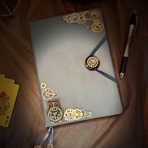 May include: A blue notebook with a steampunk design. The cover features a variety of metal gears, an owl clock, and a button closure. The notebook is lying on a brown fabric surface.