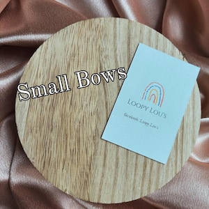 May include: A wooden round display with the text "Small Bows" in black. A small card with a rainbow design and the text "LOOPY LOU'S" is on the right side. The background is a peach-colored satin fabric.