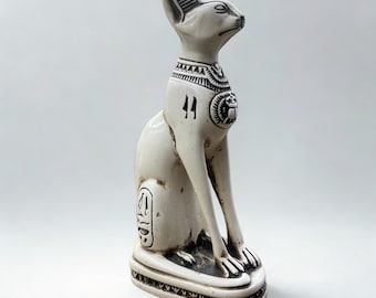 May include: A white ceramic cat statue with black detailing, in a seated pose. The statue has an Egyptian design, with hieroglyphs and a decorative collar. The cat sits on a rectangular base with a detailed border. A decorative item.