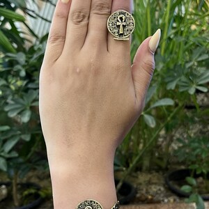 May include: A gold-toned ring and matching bracelet set featuring a circular medallion design with an embossed ankh symbol at the center, surrounded by intricate hieroglyphic-style carvings on the metal surface of both jewelry pieces.