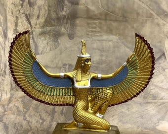 May include: A gold-coloured figurine of the Egyptian goddess Isis kneeling with large, outstretched wings. The wings feature blue and green accents with red tips. The figure wears a headdress and is mounted on a rectangular base with decorative carvings.
