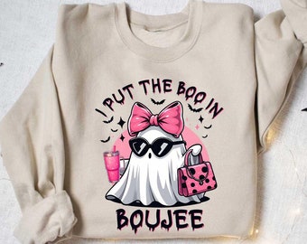 May include: A beige sweatshirt featuring a graphic of a ghost wearing sunglasses, a pink bow, and carrying a pink handbag. The text "I put the boo in boujee" is printed on the sweatshirt.
