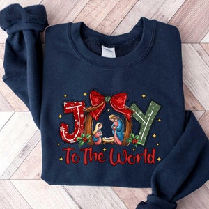 May include: Navy blue sweatshirt with a Christmas-themed design featuring the word "JOY" in red and green letters with a red bow and a nativity scene. The text "To The World" is printed below the word "JOY".