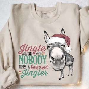 May include: Beige sweatshirt with a graphic of a donkey wearing a Santa hat. Text on the shirt reads "Jingle all the way, nobody likes a half-assed Jingler" in red and green lettering. Christmas-themed apparel.