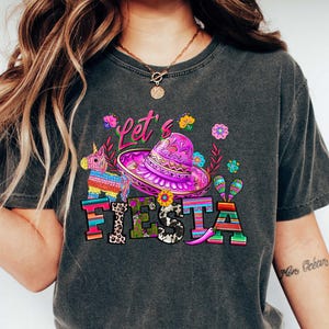 May include: Dark gray t-shirt with a colorful "Let's Fiesta" graphic. The design includes a sombrero, a piñata, maracas, and floral elements. The shirt is a casual style, suitable for festive occasions.