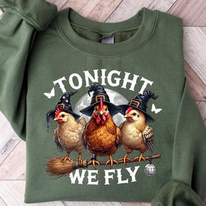 May include: An olive green sweatshirt featuring a graphic of three chickens wearing witch hats, perched on a broom. The text "TONIGHT WE FLY" is printed above the chickens. The sweatshirt has long sleeves and a crew neck.
