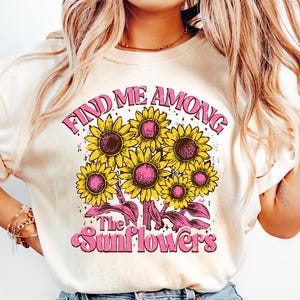 May include: A cream colored t-shirt with a graphic of sunflowers and the text "Find me among the sunflowers" in pink.
