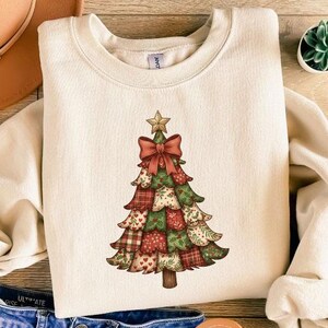 May include: Cream-colored sweatshirt featuring a Christmas tree design. The tree is adorned with a red bow, a gold star, and patchwork-style patterns in red, green, and tan. The sweatshirt is paired with blue denim shorts, a brown hat, and sandals.