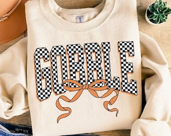 May include: Cream-colored sweatshirt with the word "GOBBLE" in a black and white checkered pattern, accented with an orange bow. The sweatshirt is paired with blue denim shorts, a tan hat, and sandals.