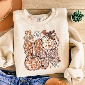 May include: Cream-colored sweatshirt featuring a fall-themed graphic. The design includes pumpkins, bows, daisies, and cherries in shades of brown, orange, and white. The sweatshirt is paired with blue denim shorts, a brown hat, and sandals.