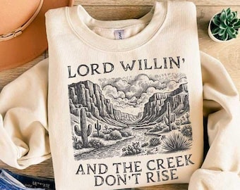 May include: A beige sweatshirt with a black and white illustration of a canyon landscape. The text "LORD WILLIN', AND THE CREEK DON'T RISE" is printed on the front of the sweatshirt.