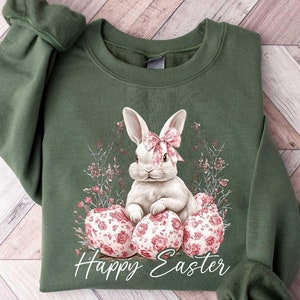May include: A green sweatshirt with a white bunny rabbit sitting on pink and white floral Easter eggs. The text "Happy Easter" is printed below the bunny.