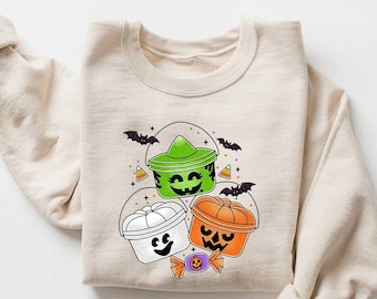 Könnte beinhalten: Ein beiger Pullover mit einem Halloween-Motiv, das drei Eimer mit Kürbisgesichtern zeigt. Die Eimer sind grün, weiß und orange. Das Design enthält auch Fledermäuse und Maisbonbons.