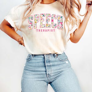 May include: A light yellow t-shirt with a floral design spelling out the word "SPEECH" and the text "THERAPIST" underneath.