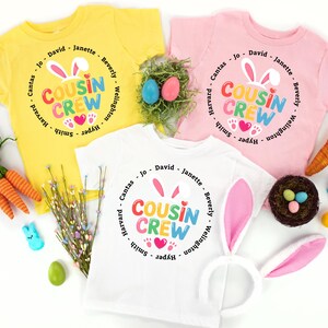 May include: Three children's t-shirts in yellow, pink, and white, each printed with "COUSIN CREW" in a circle design with bunny ears and hearts. Easter decorations include carrots, eggs, and a bunny ear headband.
